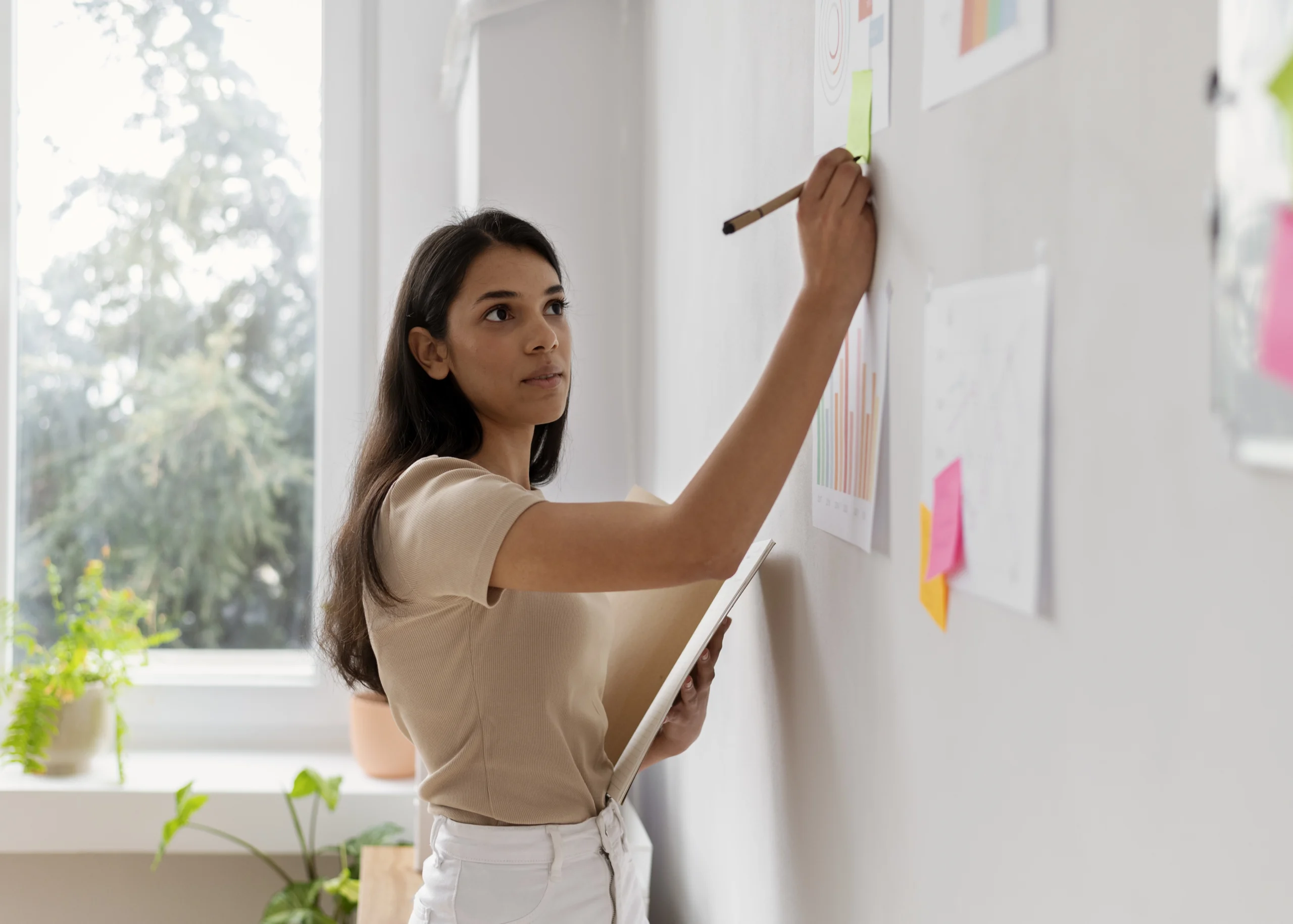 Mulher jovem escreve em post-it em uma parede com gráficos e anotações, segurando um caderno.